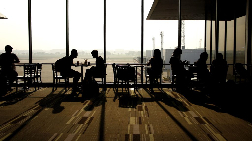 believable characters: several silhouetted people sit at tables in front of windows in an airport (coincidentally the best place to people-watch for characters!)